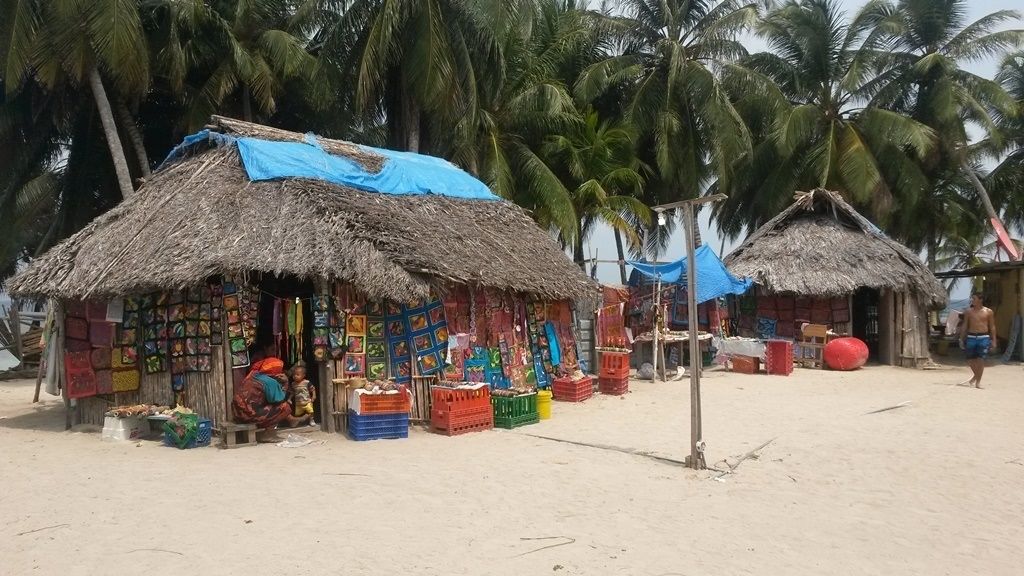 Un marché installé au bord de la plage, avec des étals aux toits de chaume où sont vendus des articles aux couleurs vives.