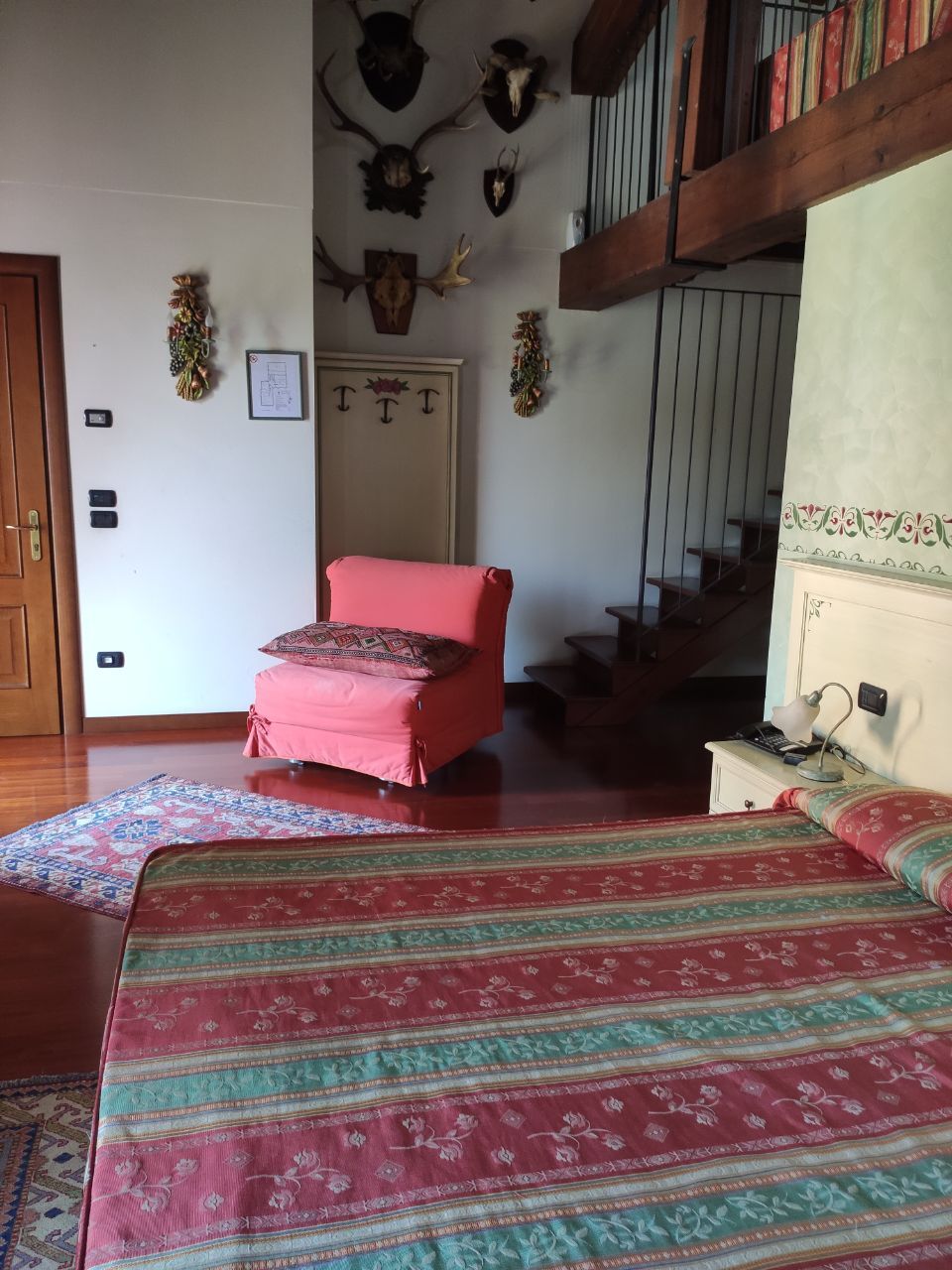 A room with a bed, a chair, a staircase, and wall-mounted deer antlers.