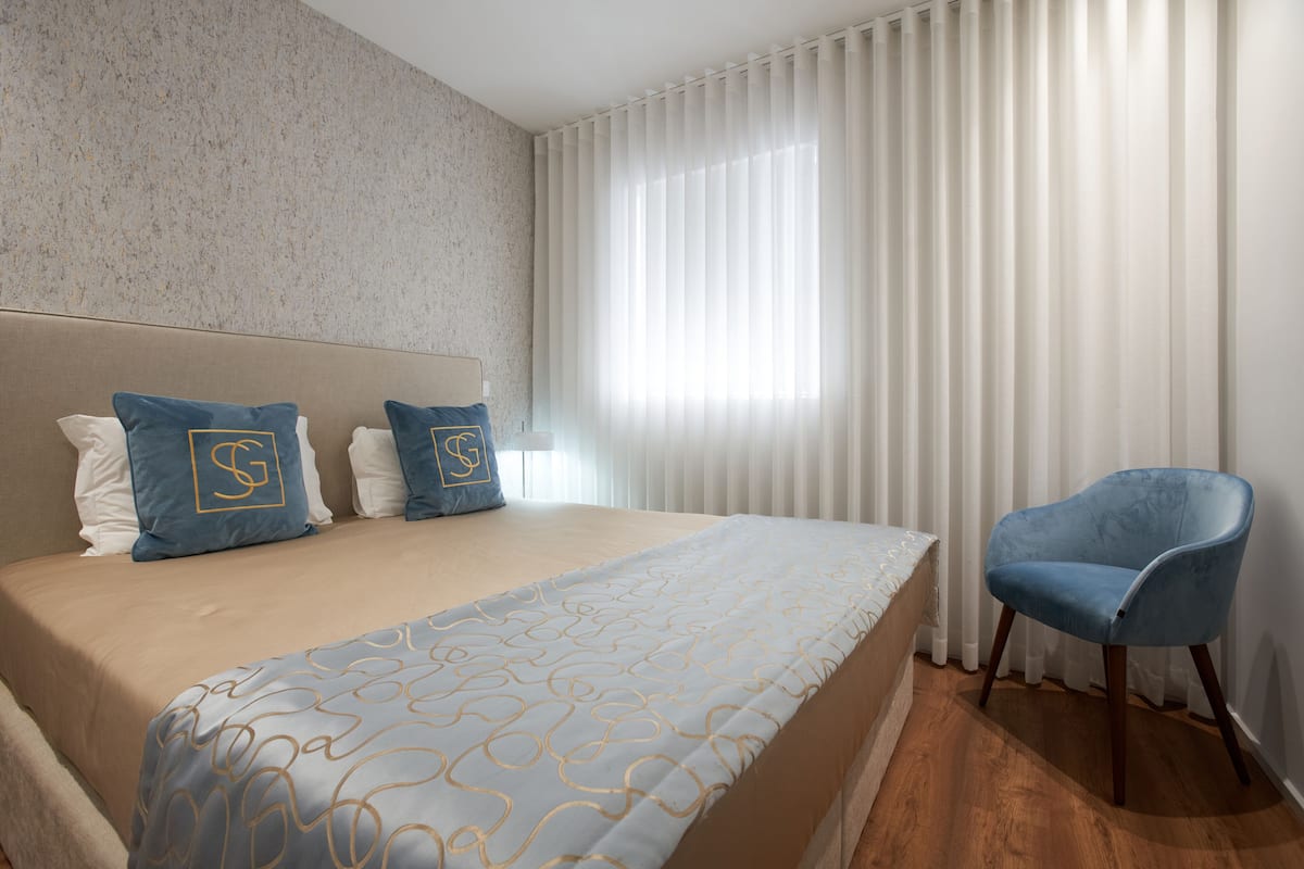 A hotel room with a large bed, a blue chair, and a window with white curtains.