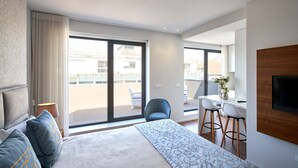 Quarto de hotel moderno com uma cama grande, uma varanda com vista, um espaço de refeições com duas cadeiras e uma TV fixada na parede.