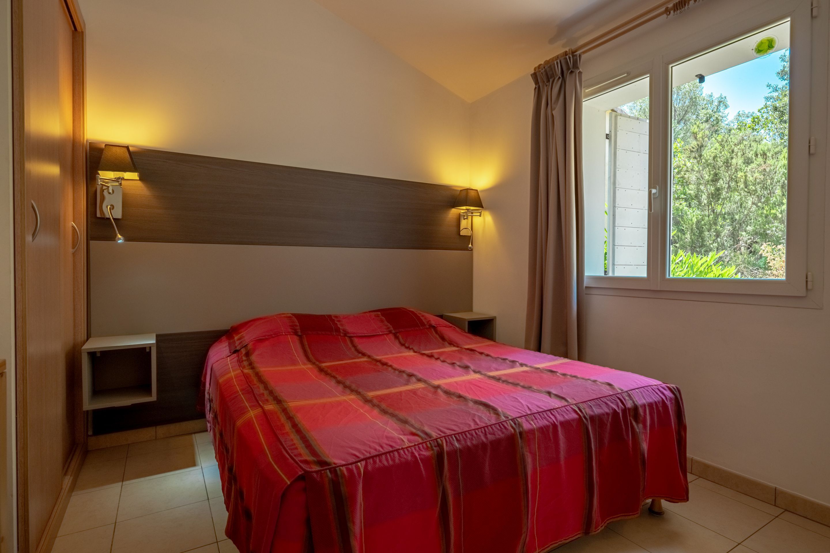 Un dormitorio con cama, dos lámparas de noche, una ventana con cortinas y una pequeña estantería.
