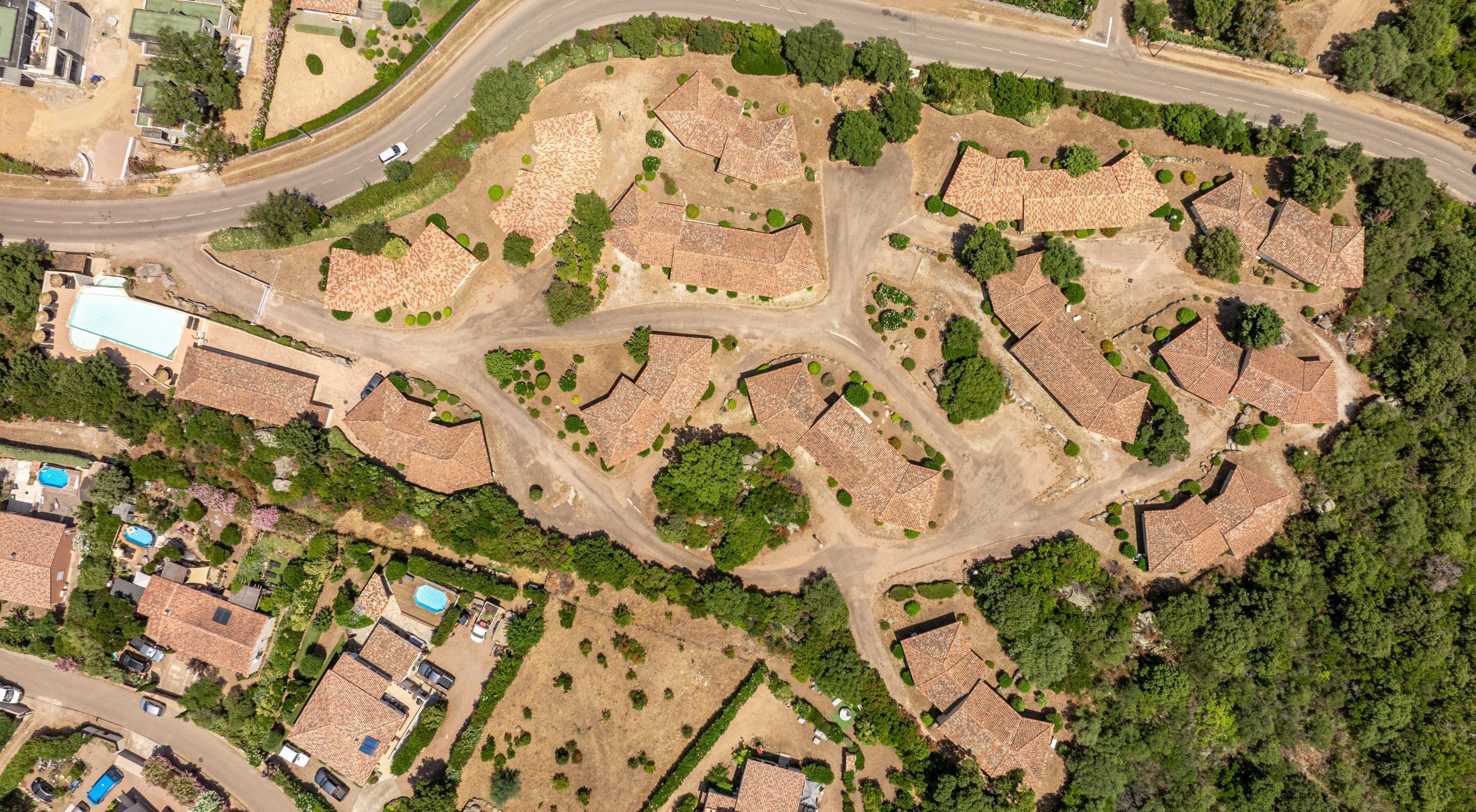 An aerial view of a residential area with terracotta-roofed houses, a winding road, and surrounding greenery.