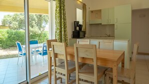 A dining area with a wooden table and chairs, a kitchen with light green cabinets, and a sliding glass door leading to a patio with blue chairs and greenery. - Résidence Odalys San Martinu (Zonza)