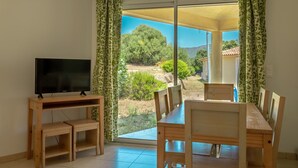 A room with a TV on a wooden stand, a dining table with chairs, and a sliding glass door leading to a garden with trees and a house in the background. - Résidence Odalys San Martinu (Zonza)