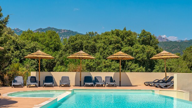 A pool area with lounge chairs and thatched umbrellas, surrounded by lush greenery and mountains in the background. - Résidence Odalys San Martinu (Zonza)