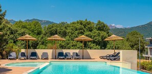 A pool area with lounge chairs and thatched umbrellas, surrounded by lush greenery and mountains in the background. - Résidence Odalys San Martinu (Zonza)