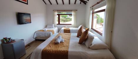 A hotel room with two beds, a television, a small desk, and a window with a view of greenery.