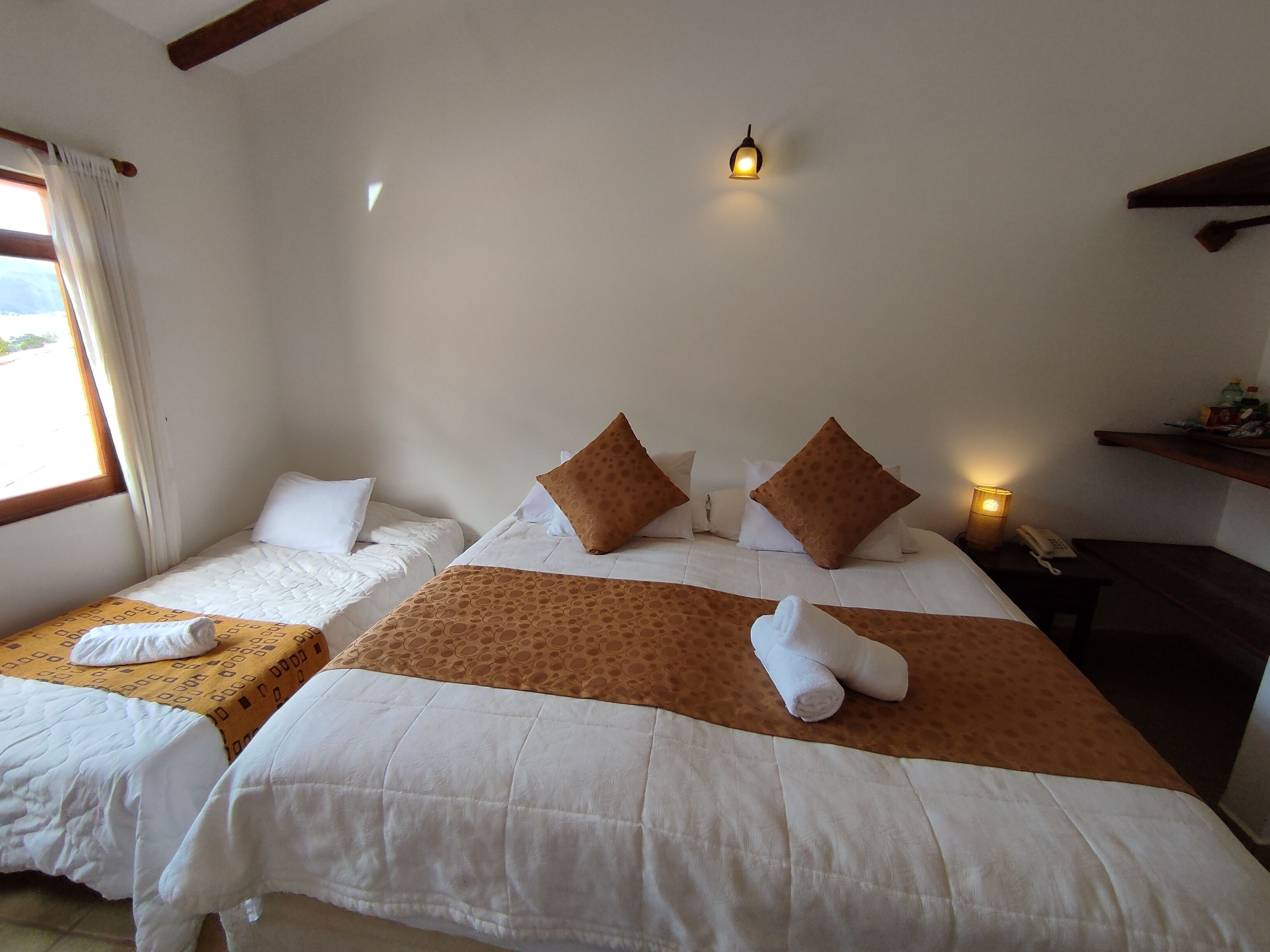 A hotel room with two beds, each with white bedding and brown decorative pillows. There is a small table with a lamp and a phone on it.
