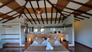 Un dormitorio espacioso con un techo de madera alto, una cama grande con cabecera de madera, mesitas de noche con lámparas y vistas a un baño con lavamanos y espejo.