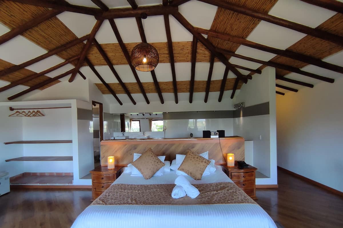 Une chambre spacieuse avec un haut plafond en bois, un grand lit doté d’une tête de lit en bois, des tables de chevet avec des lampes, et une vue sur une salle de bain équipée d’un lavabo et d’un miroir.