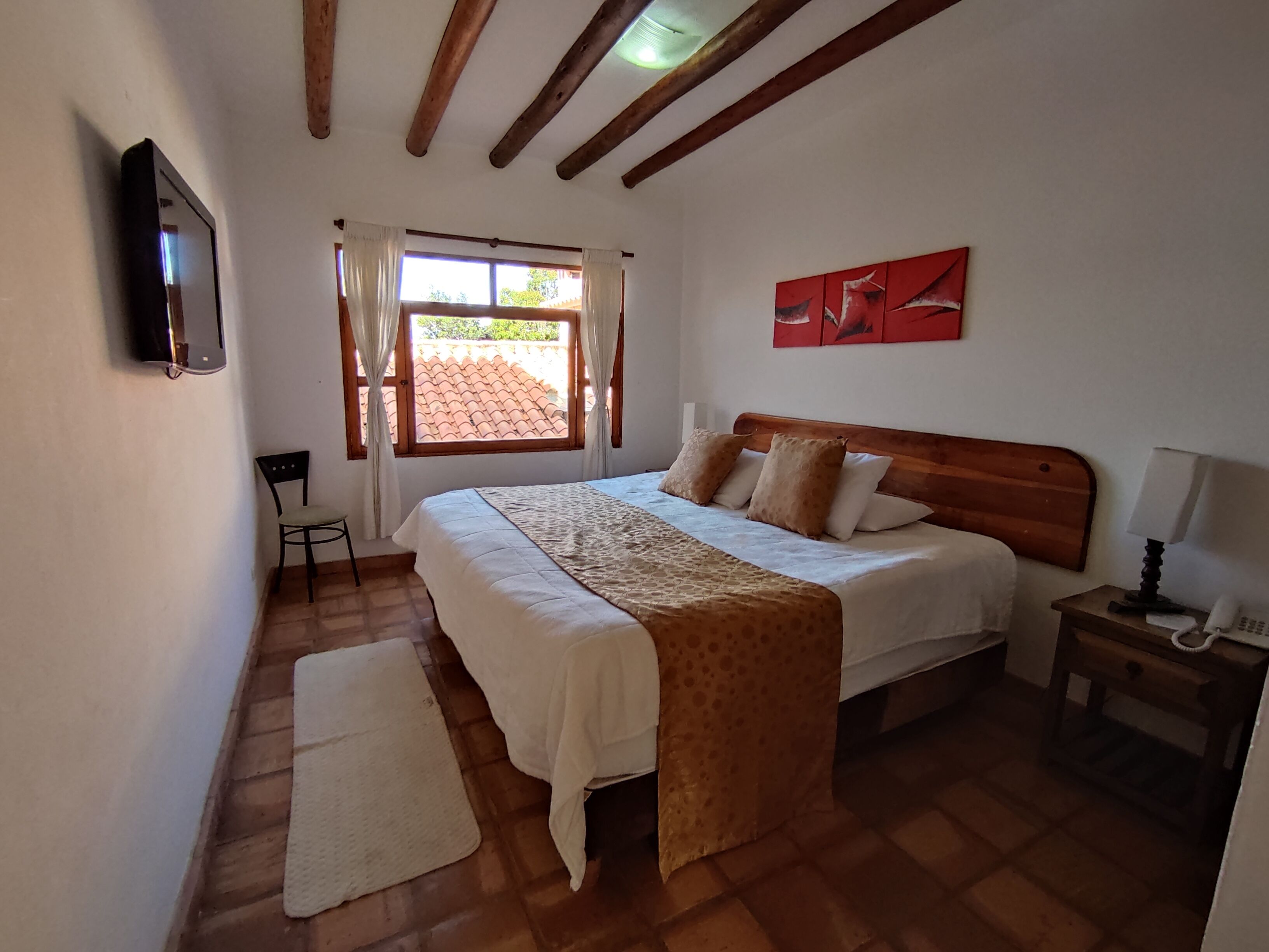 A bedroom with a bed, a nightstand, a chair, a window with curtains, and a wall-mounted TV.