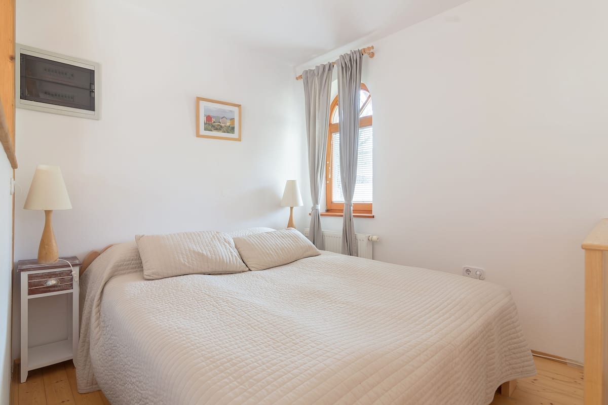 A bedroom with a bed, bedside tables, a window with curtains, a wall-mounted air conditioning unit, and a framed picture.