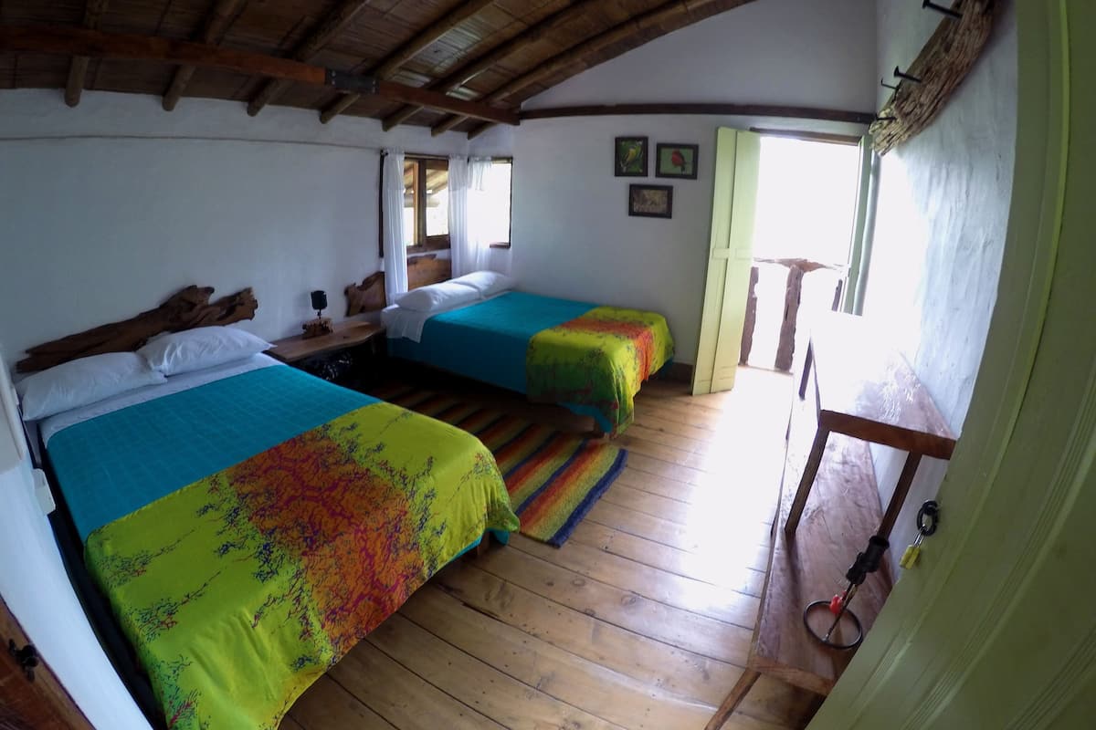 A room with two beds, wooden flooring, a rustic ceiling, and a window with curtains.