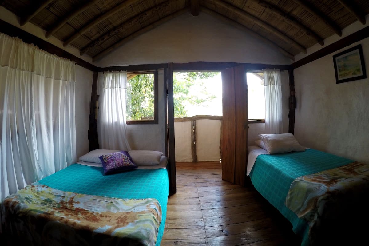 A room with two beds, wooden flooring, a sloped ceiling, and a window with curtains.
