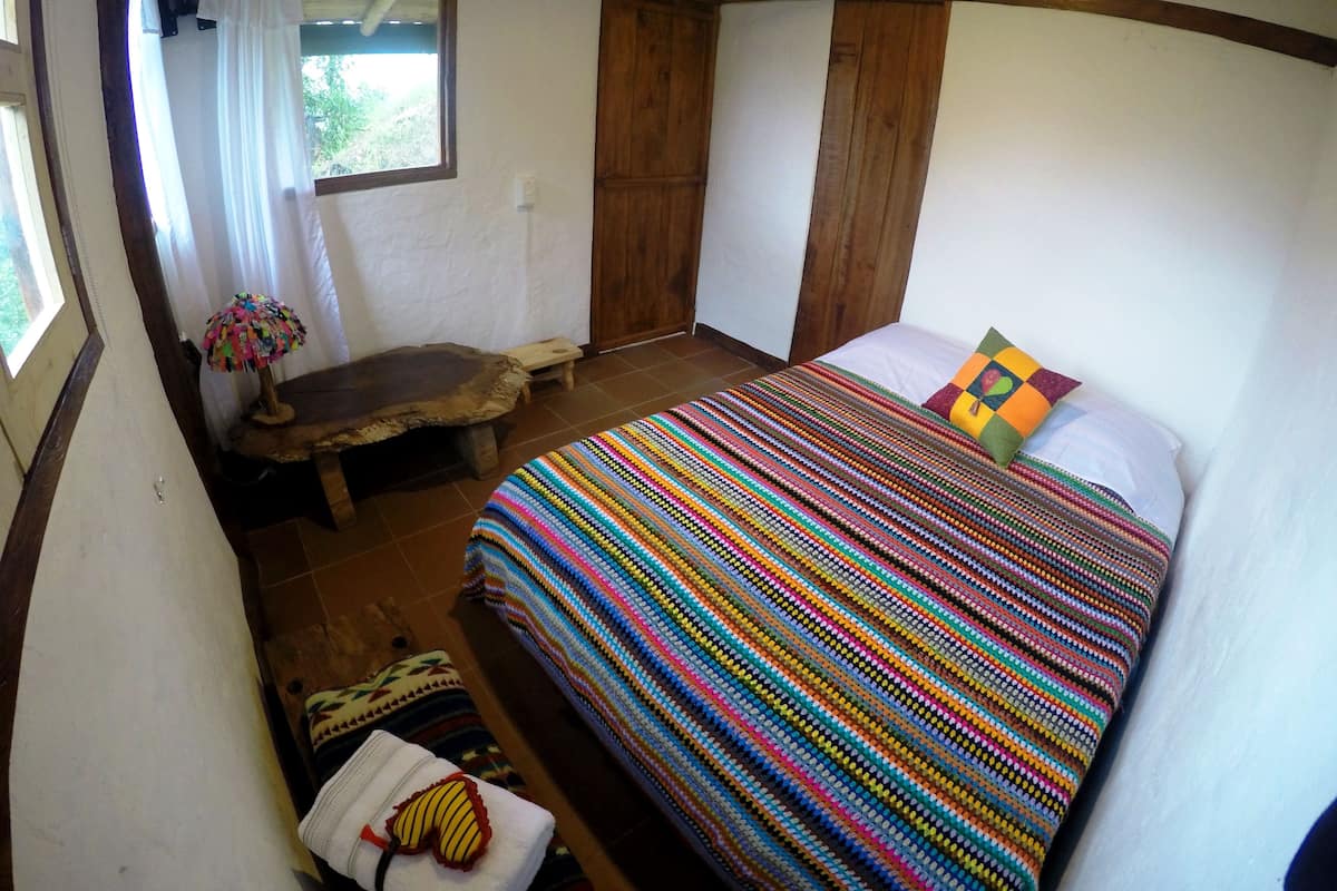A bedroom with a wooden bed, a colorful blanket, a small bench, and a window with curtains.
