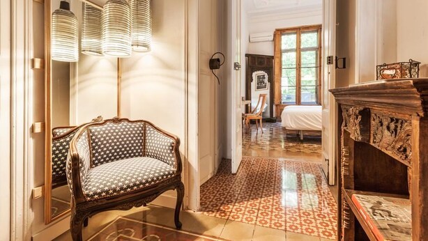 A room with a patterned floor, a wooden chair, a mirror, and a bed with a window in the background. - Factory Suites Barcelona (Barcelona)