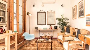 A cozy living room with a wooden coffee table, a blue chair, and a sofa. There is a small desk with a lamp and a plant. - Factory Suites Barcelona (Barcelona)