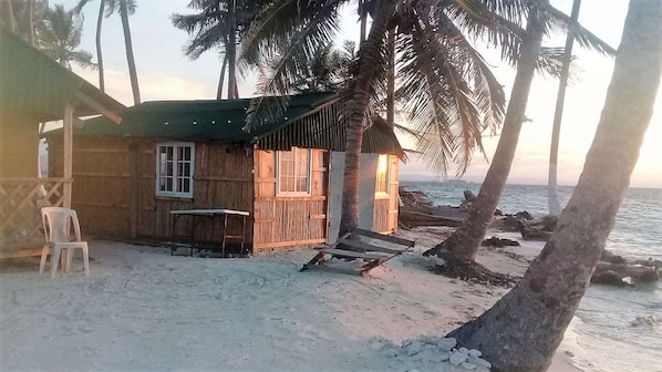 A wooden beach hut with a table and chairs, a window, and a view of the ocean.
