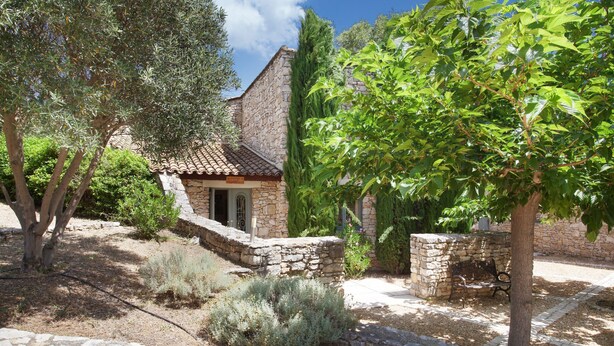 A stone building with a tiled roof, surrounded by olive trees and other vegetation.