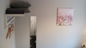 A bedroom with a bed, a bedside table, a mirror, a wall-mounted shelf with a pillow, a painting, and a door leading to a bathroom. - Templemore Town House (Belfast)