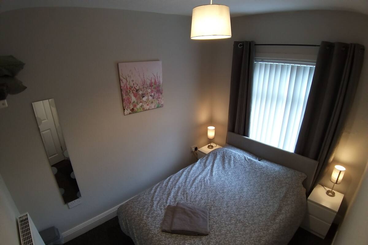 A bedroom with a bed, bedside lamps, a window with curtains, a wall-mounted painting, and a ceiling light.