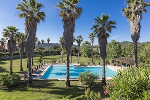 A swimming pool surrounded by palm trees and a grassy area with a view of hills in the background. - Résidence Odalys Stella di l'Aria (Sartene)