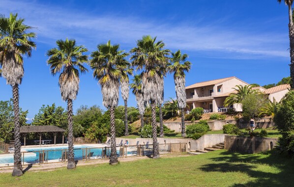 A pool area with palm trees, a gazebo, and a building with a red roof. - Résidence Odalys Stella di l'Aria (Sartene)
