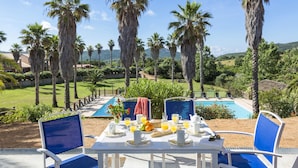 Une table à manger avec des chaises, des aliments pour le petit-déjeuner, et une vue sur une piscine et des palmiers.