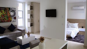 A modern living room with a sofa, coffee table, and artwork. A bedroom with a bed and TV is visible in the background. - Loft Mikonos - Adults Only (Pereira)