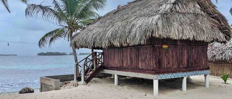 A thatched-roof hut with a wooden deck and a small staircase leading up to it, situated on a sandy beach with palm trees and a view of the sea.