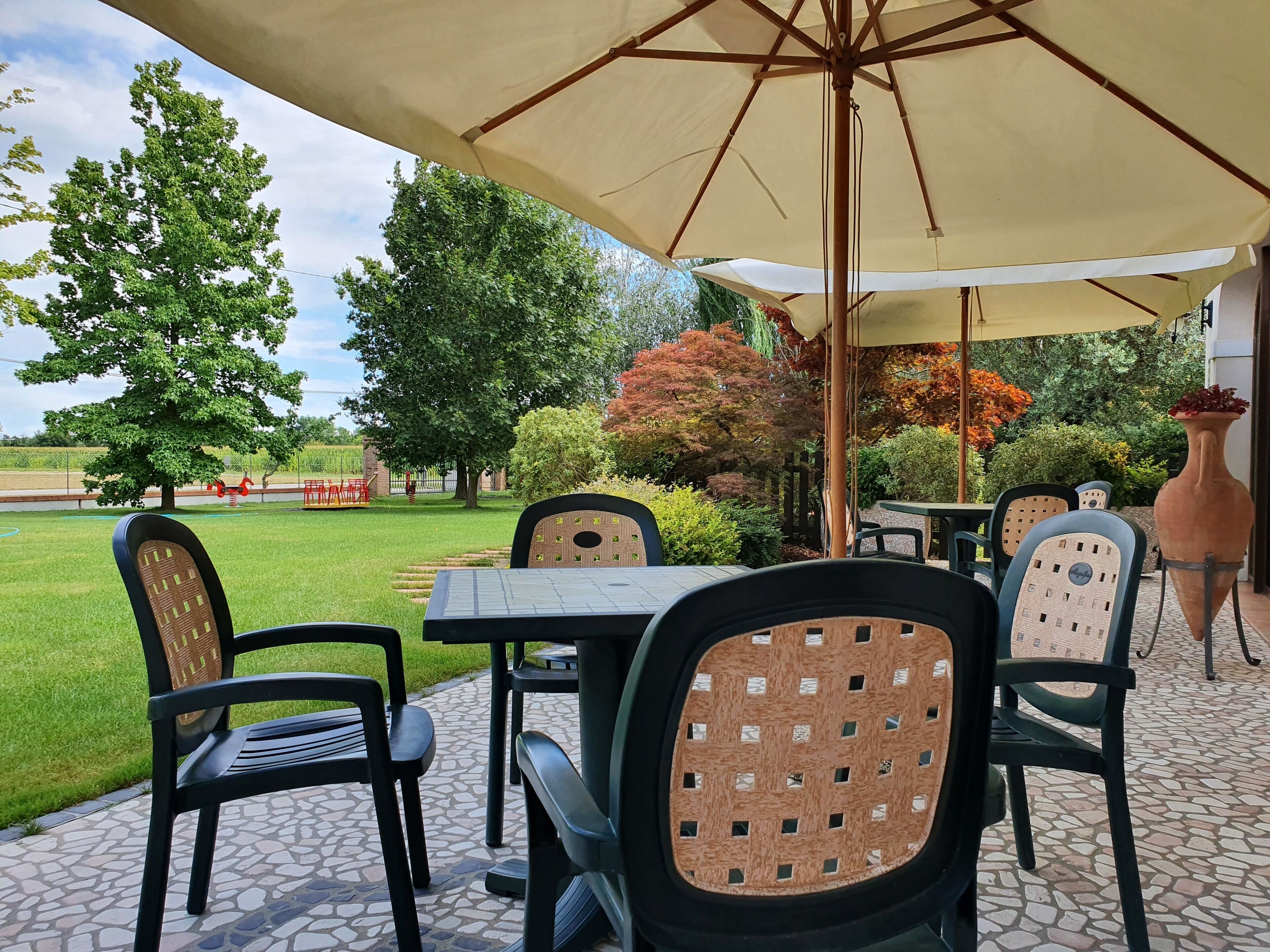 An outdoor patio area with a table and chairs under a large umbrella, surrounded by greenery and trees.