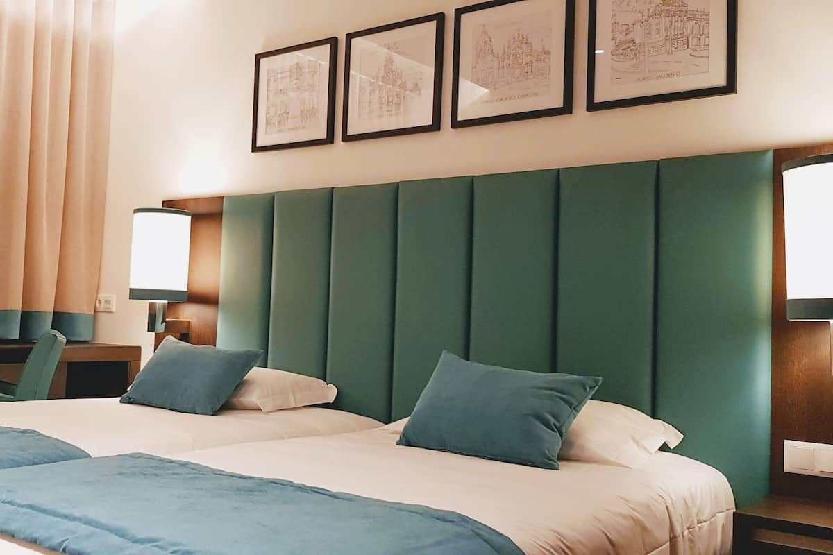 A hotel room with two beds, a green padded headboard, a desk, and a window with curtains.