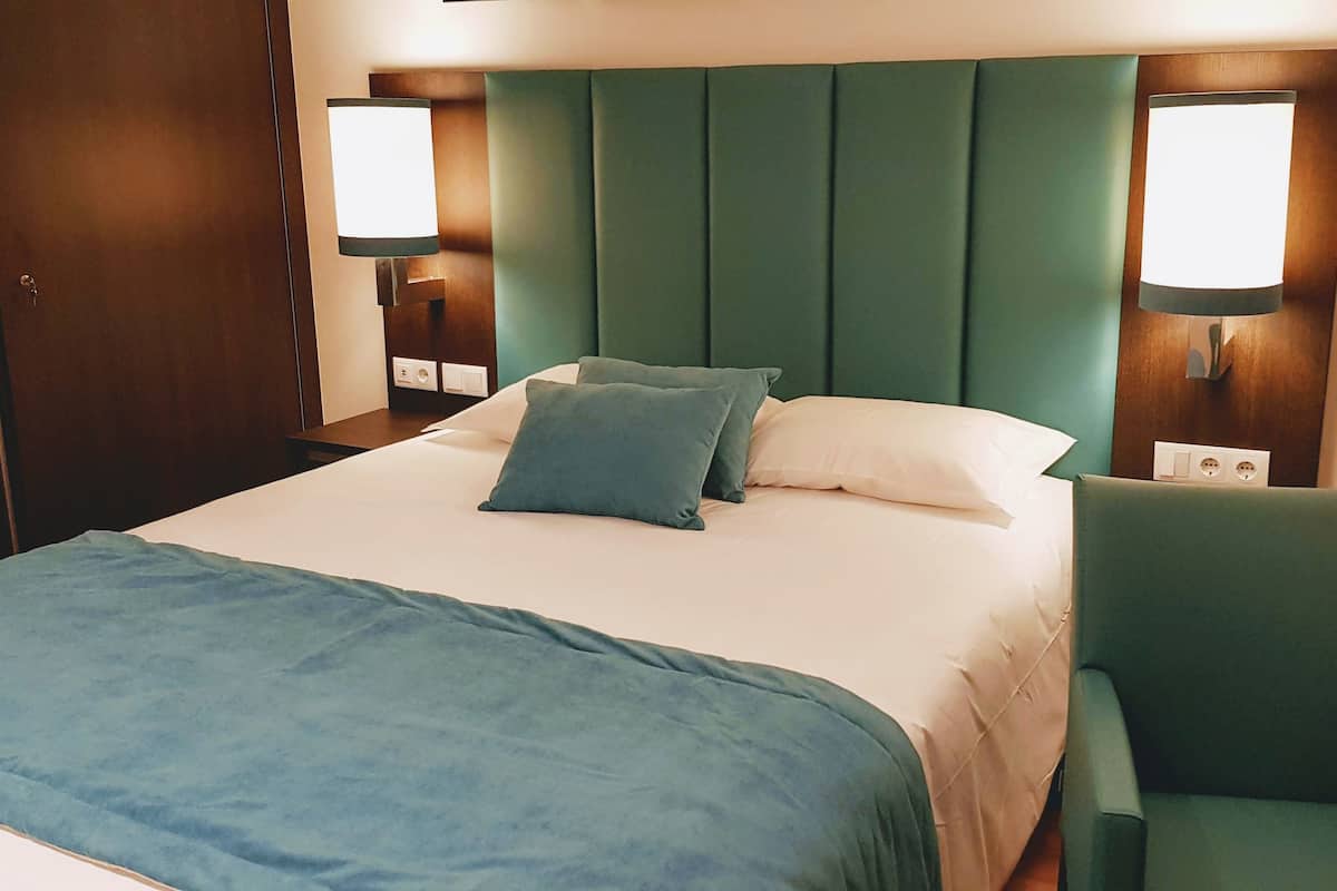 A neatly made bed with a green headboard, two pillows, and a blue throw blanket. There are two wall-mounted lamps on either side of the bed.