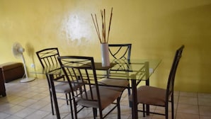 A dining area with a glass table, four chairs, a white vase with sticks, and a standing fan. - Tapion Beach Suites 1 (Castries)