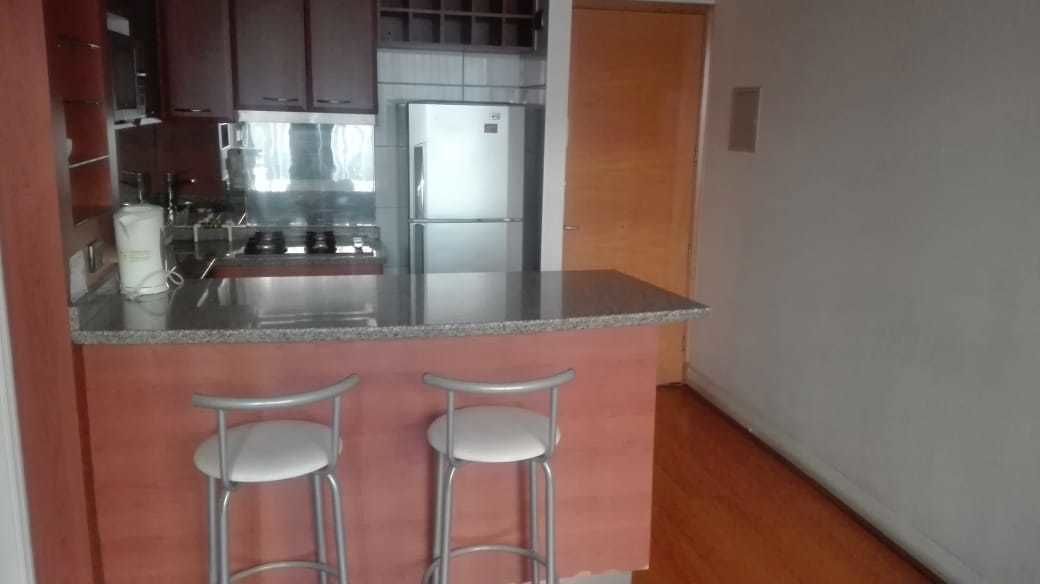 A compact kitchen with a stainless steel refrigerator, a built-in oven, and a granite countertop. There are two bar stools with white seats.