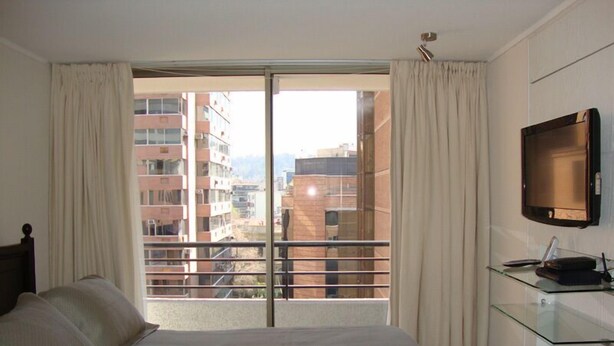Quarto com cama grande, televisão na parede e varanda com vista para edifícios.