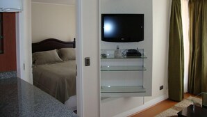 Quarto de hotel com uma cama, uma televisão fixada numa prateleira de vidro e uma porta que dá para outro quarto.