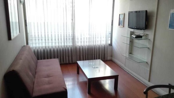 A living room with a sofa, a coffee table, a TV mounted on the wall, and a glass shelf unit.