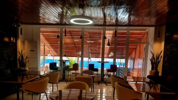 Un restaurante con vista al mar, con mesas y sillas redondas, techo de madera y grandes ventanales.