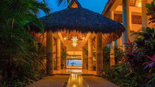 A resort entrance with a thatched roof, palm trees, and a view of the ocean.