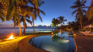 A pool surrounded by palm trees and lounge chairs, with a view of the ocean at dusk.