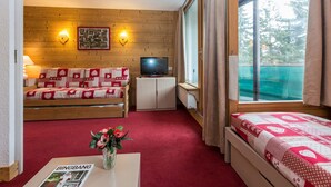 A cozy room with a wooden wall, a sofa with a red and white patterned cover, a television on a cabinet, a bed with a red and white patterned cover, and a window with curtains. - Résidence Odalys Chanteneige Croisette (Les Belleville)