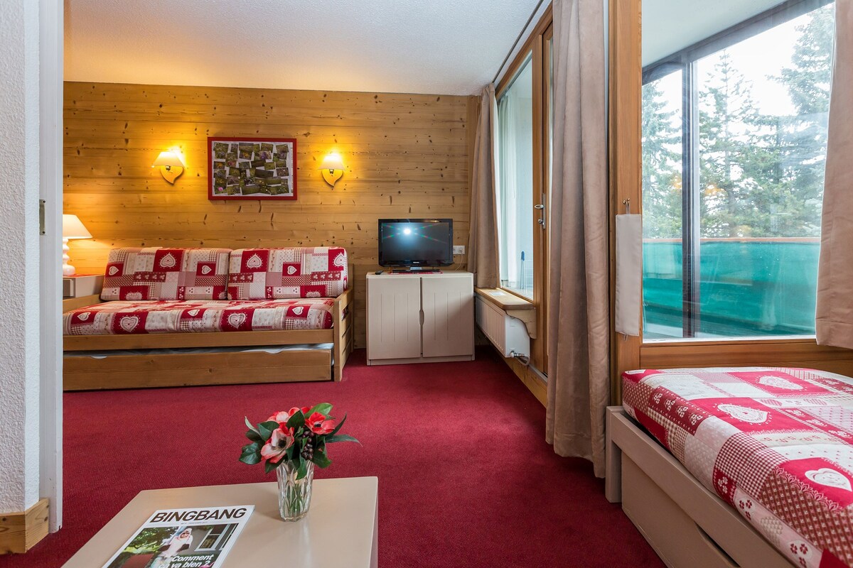 Quarto com parede de madeira, um sofá com capa estampada em vermelho e branco, uma televisão em um móvel, uma janela com cortinas e uma cama com capa estampada em vermelho e branco.