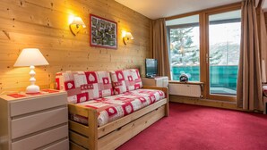 A cozy room with a wooden wall, a red carpet, a sofa with a quilt, a TV, a lamp, and a window with curtains. - Résidence Odalys Chanteneige Croisette (Les Belleville)