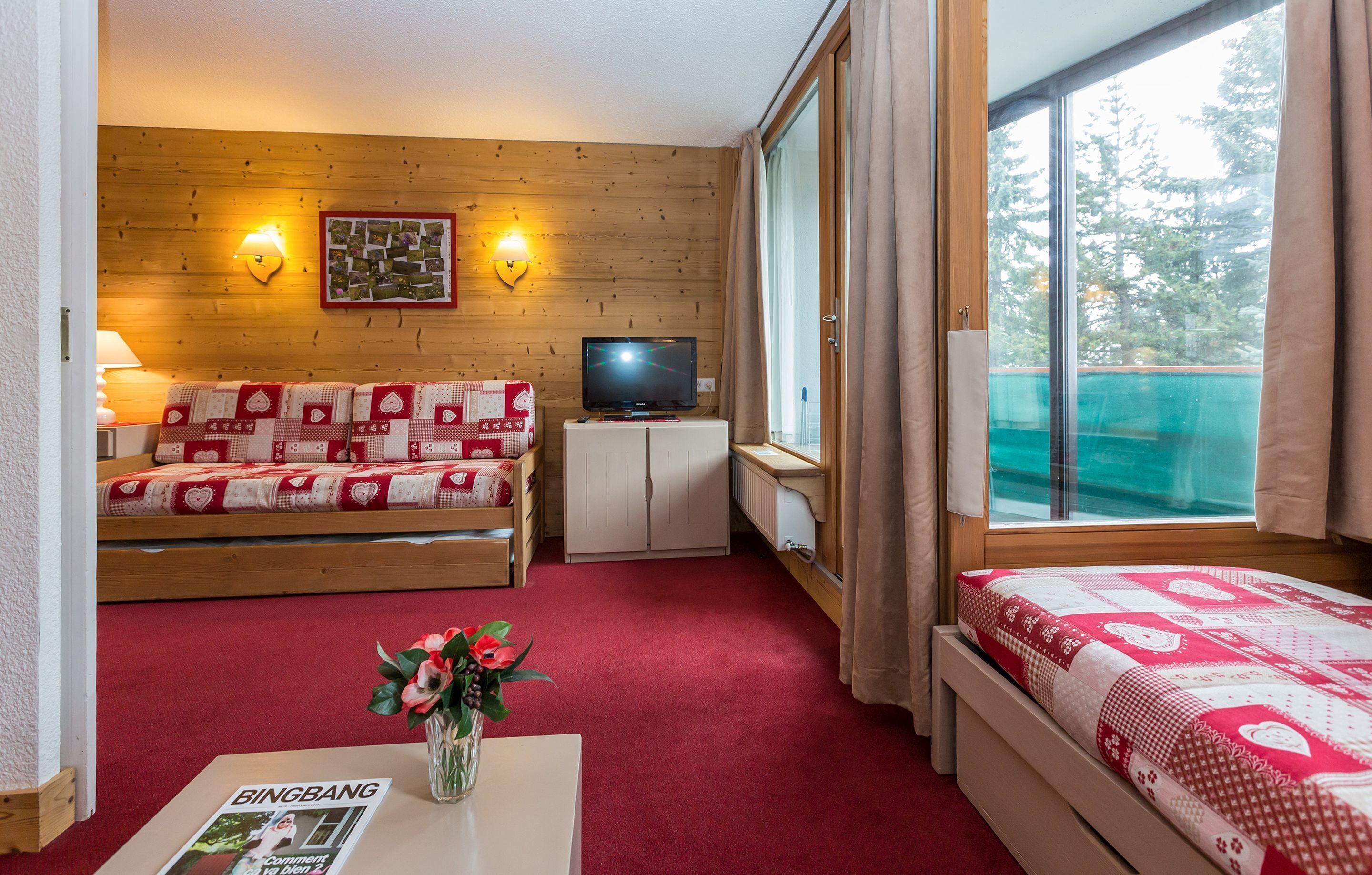 A room with a wooden wall, a sofa with a red and white patterned cover, a television on a cabinet, a window with curtains, and a bed with a red and white patterned cover.