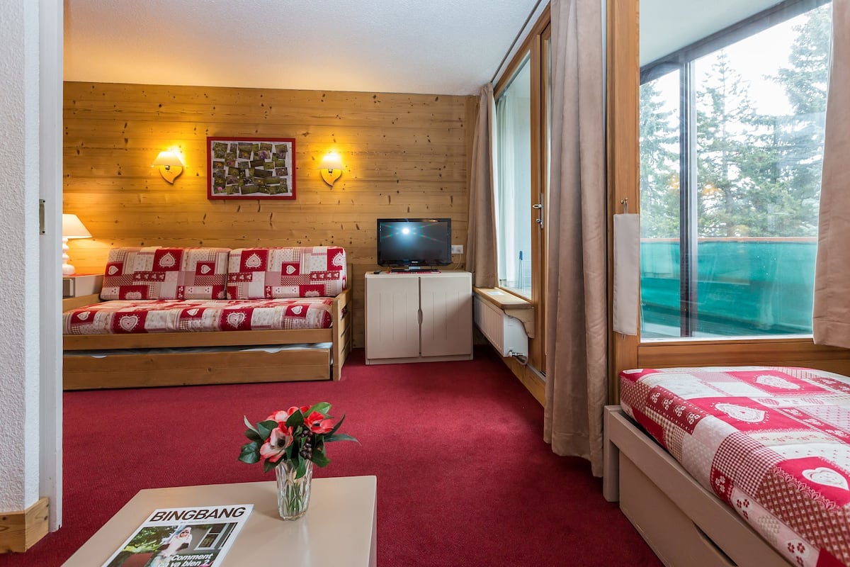A room with a wooden wall, a sofa with a red and white patterned cover, a television on a cabinet, a window with curtains, and a bed with a red and white patterned cover.
