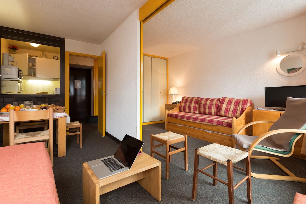A compact hotel room with a kitchenette, a sofa, a chair, a small table with a laptop, and a TV.