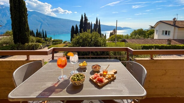 Una mesa con vistas a montañas y un lago, un vaso de zumo de naranja, un bol de aceitunas y un plato con queso y jamón.