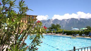 Una piscina con sillas de descanso y sombrillas, rodeada de montañas y un edificio.