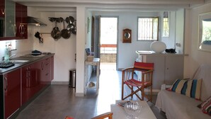 Amplia sala de estar con sofá, mesa de centro y mesa de comedor. Se observa una cocina con armarios rojos y estufa. Hay una ventana con rejas y una silla ubicada cerca de la ventana.